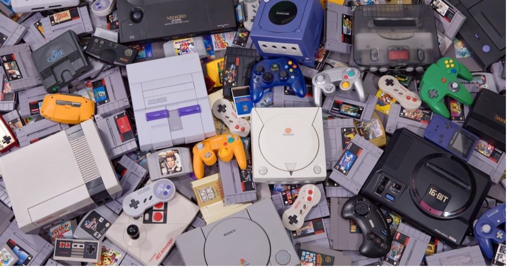 A flat-lay of classic retro video game consoles, controllers, and cartridges scattered together, including Super Nintendo, Nintendo 64, Sega Genesis, PlayStation, Dreamcast, GameCube, and Neo Geo systems.