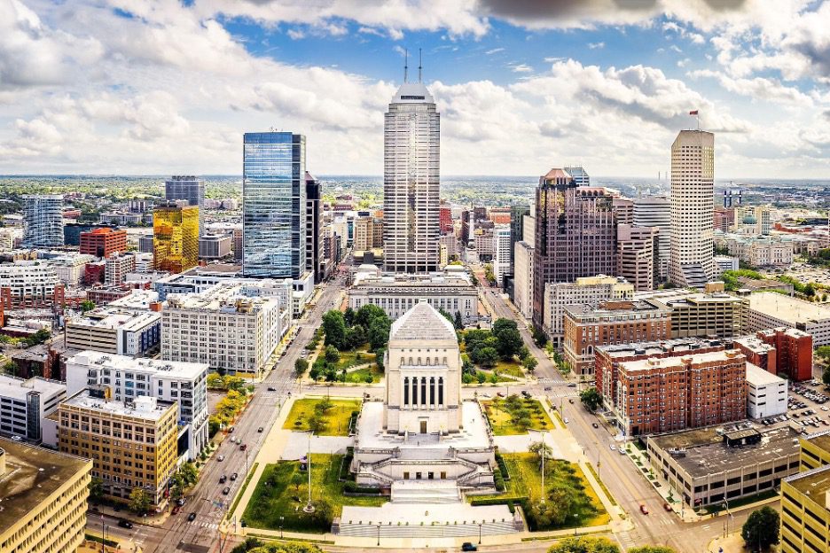 Aerial view of downtown Indianapolis featuring Monument Circle and surrounding skyscrapers under a partly cloudy sky.