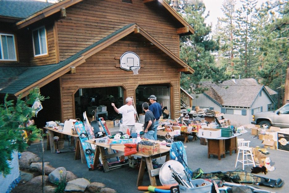 A residential garage sale with tables set up in a driveway, people browsing assorted household items, electronics, and boxes outside a suburban home.