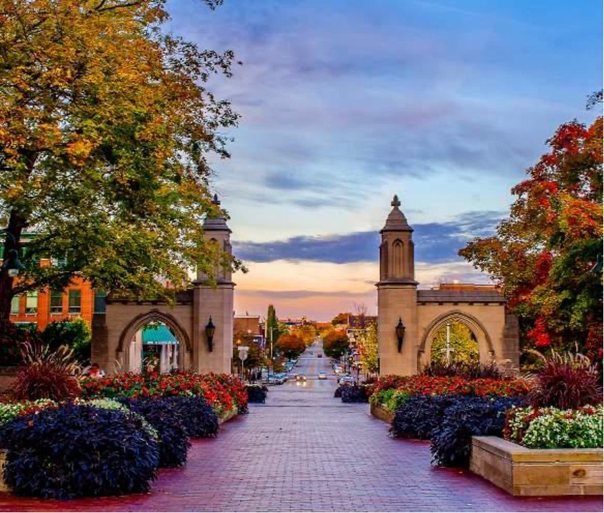 Autumn view of the Sample Gates at Indiana University in Bloomington, with colorful fall foliage, landscaped flower beds, and a brick walkway leading toward downtown at sunset.